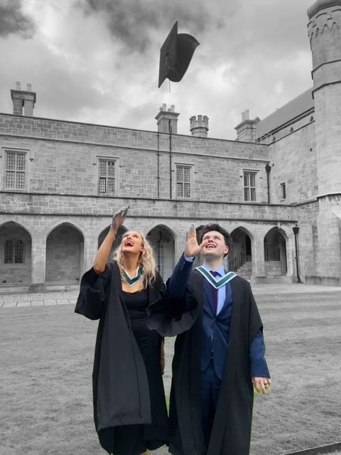 Two graduates in gowns throwing caps in the air outside a historic stone building.